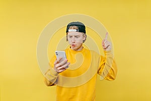 Surprised guy in a cap and colored sweatshirt uses a smartphone and points his finger up at the copy space, with a shocked face