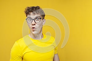 Surprised guy student with curly hair holding glasses and looking at copy space, nerd in glasses on yellow background