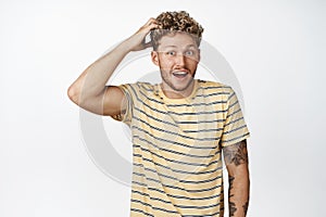 Surprised handsome guy smiling, awkwardly scratching his head, looking puzzled and happy, standing over white background