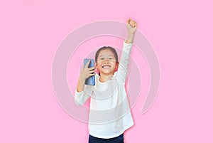 Surprised happy asian little child girl watching her smartphone and expression hand fight sign over pink background with copy