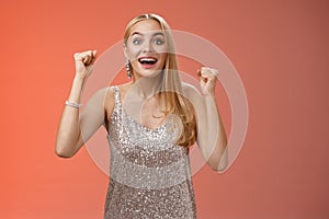 Surprised happy celebrating blond young woman in silver trendy dress raising hands up yes victory gesture smiling