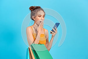 Surprised happy woman holds cheap paper shopping bags looks at phone screen read sms sell out promotion store isolated on blue