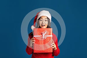 Surprised happy woman holds in hands christmas gift. Beautiful woman in santa claus hat on blue background Xmas concept