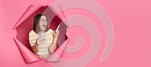 Surprised happy woman wearing striped shirt breaking through pink paper hole holding samrtphone pointing away at copy space for