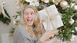 Surprised and happy young woman with christmas present box near christmas tree.