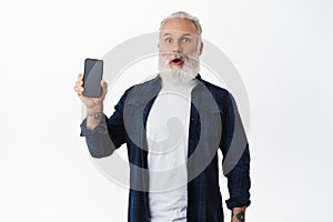 Surprised and impressed senior man showing mobile phone screen, holding smartphone in hand, gasping and looking amazed