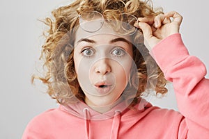 Surprised intrigued curly-haired feminine woman taking off glasses and saying wow while standing over gray background