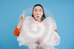 Surprised korean woman with plastic waste, points finger up and looks on top, blue background