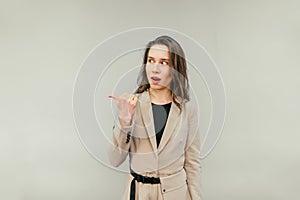 Surprised lady in a beige suit shows her finger aside on copy space and looks with a shocked face, isolated on a beige background