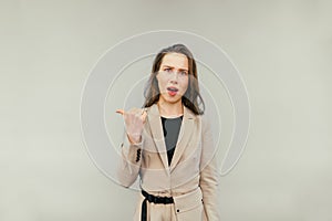 Surprised lady in a beige suit stands on a beige background, looks into the camera with a dissatisfied face and points a finger to