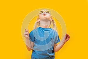 Surprised little blonde girl with glasses looks up. Studio portrait of child in blue T-shirt on yellow background