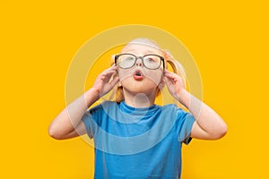 Surprised little girl adjusts her glasses and looking up. Portrait of little blonde girl in blue T-shirt with glasses on yellow