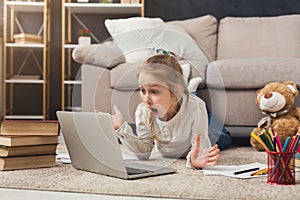 Surprised little girl doing homework on laptop, lying on floor