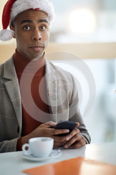 Surprised look of young afro-american in the office, texting on cell phone, wearing santa hat with cup of coffee in front of him