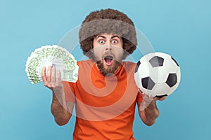 Surprised man with Afro hairstyle holding soccer ball and fan of euro bills, sports betting, big win