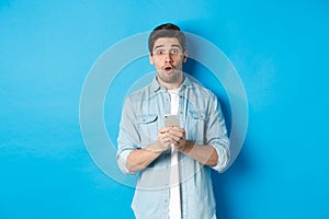 Surprised man in casual clothes looking astounded, holding smartphone, standing against blue background