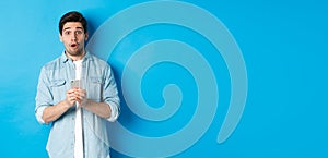 Surprised man in casual clothes looking astounded, holding smartphone, standing against blue background