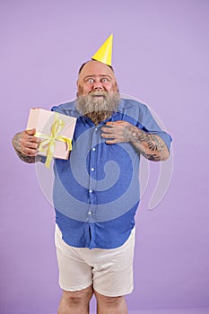 Surprised man with overweight in tight blue shirt holds gift box posing on purple background