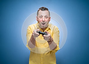 Surprised man playing video games with joystick over blue background, dresses in yellow shirt. Astonished guy holding game