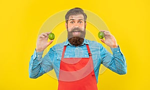 Surprised man in red apron holding fresh limes citrus fruits yellow background, fruiterer photo