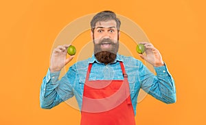 Surprised man in red apron holding fresh limes citrus fruits yellow background, fruiterer