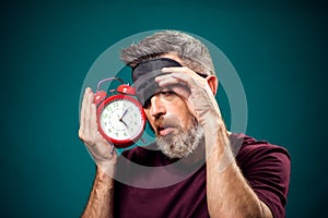Surprised man in red t-shirt and sleep mask on head holding alarm clock and pillow. Lifestyle and bed time concept
