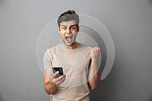 Surprised man with short dark hair shouting and holding smartphone with clenching fist, isolated over gray background