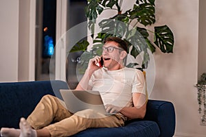 Surprised middle aged man talking on mobile phone lying on couch with laptop
