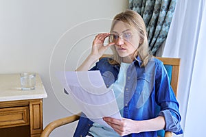 Surprised middle aged woman with glasses reading papers sitting at home in chair