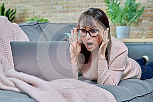 Surprised middle aged woman lying at home on sofa looking at laptop screen