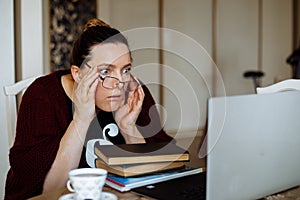 Surprised middle-aged woman sitting at table near books, looking at laptop, adjusting glasses, not believing eyes.