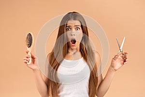 Surprised millennial girl with long straight hair holding scissors and brush on beige background