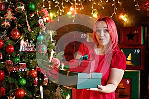 A surprised model girl with pink hair and a dress holding a gift box and posing near a Christmas tree. A young beautiful woman