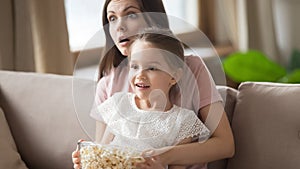 Surprised mother with little daughter watching tv, eating popcorn