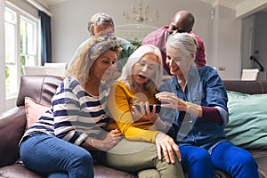 Surprised multiracial senior female friends looking at smart phone while men peeking from behind