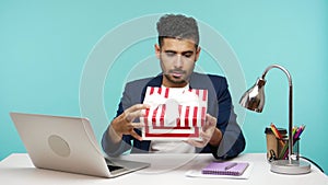 Surprised nosy business worker looking inside gift box sitting at his workplace, disappointed unsatisfied with present