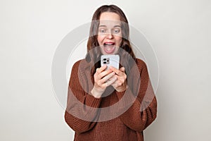 Surprised overjoyed brown haired adult woman wearing brown sweater posing isolated over gray background exclaiming with happiness