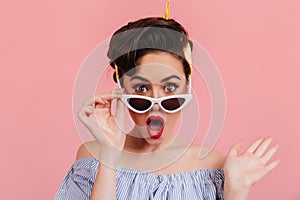 Surprised pinup girl with red lips touching sunglasses. Studio shot of amazed elegant woman isolated on pink background