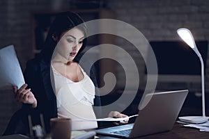 Surprised pregnant girl examines documents behind laptop. Pregnant in dark office.