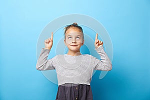 Surprised pretty little girl pointing with two fingers to up, looking to up, isolated on blue