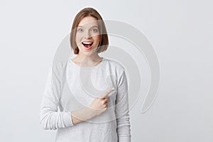 Surprised pretty young woman in longsleeve with opened mouth smiling and pointing to the side isolated over white background Feels