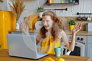 A surprised red-haired woman using a laptop at home in the kitchen. A student girl is working on a computer. Work at home,