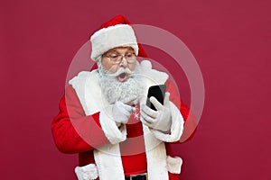 Surprised Santa Claus using smart phone isolated on red background.