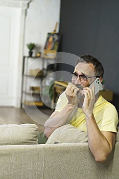 Surprised senior man wearing glasses sitting in living room experiencing astonishment when talking over smartphone