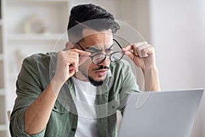 Surprised serious young arab man with beard takes off his glasses looks at laptop in home office interior