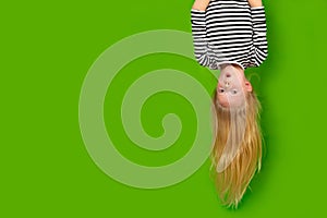 Surprised shocked little blonde girl hanging happy upside down over isolated green studio background. Emotion