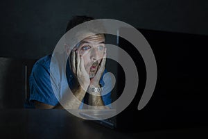 Surprised and shocked man at night working with laptop computer in the dark in disbelief and surprise face expression watching