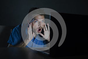 Surprised and shocked man at night working with laptop computer in the dark in disbelief and surprise face expression watching