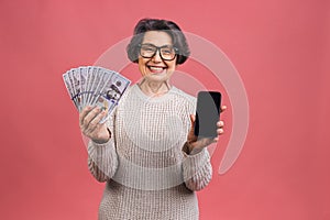 Surprised smiling happy senior mature aged senior woman in casual showing blank smartphone screen isolated over pink background.