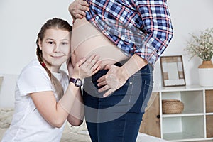 Surprised teenage daughter listening baby in belly of her pregnant mother and touching with hands, girl looking at camera